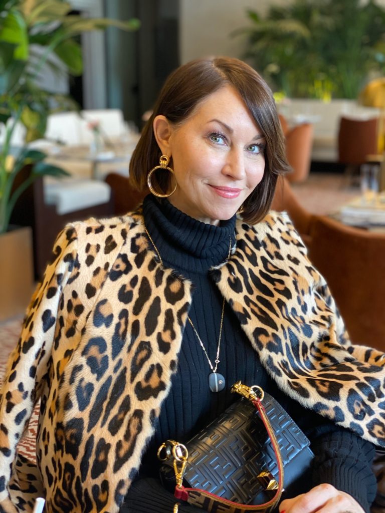 Woman wearing a big hoop earring, sitting down wearing a leopard coat and black turtleneck, holding a purse
