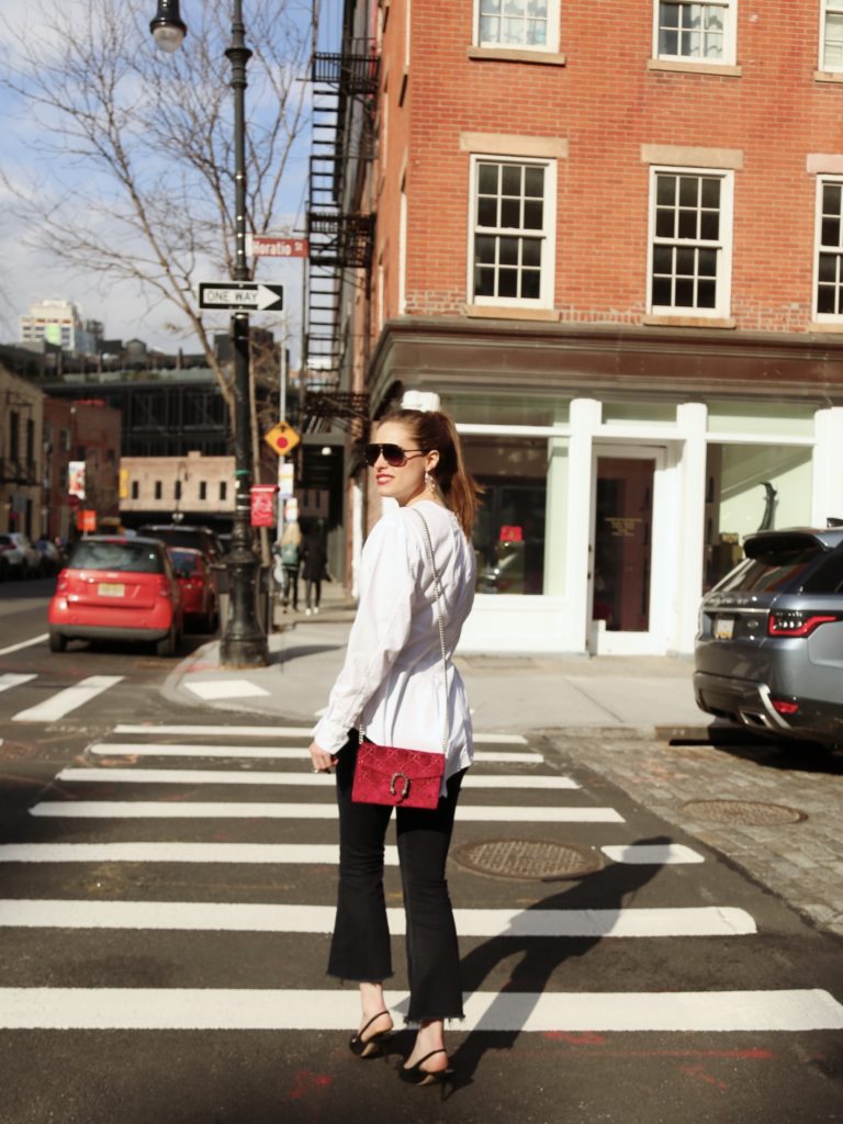 Woman walking across the street in the west village of Manhattan wearing black sunglasses, a white button-down top, black jeans and heels