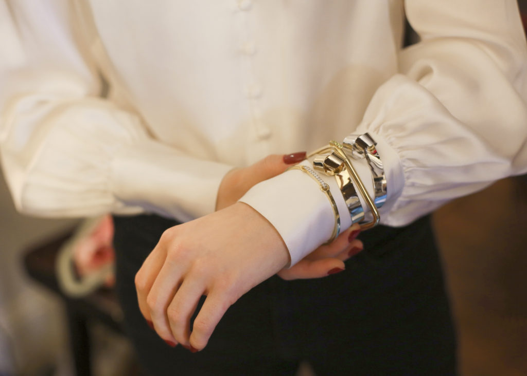 Close up photo of a woman wearing a white silk top with multiple bracelets stacked on her arm