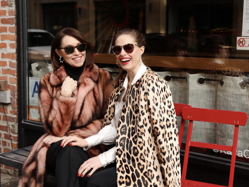 Two women sitting on red chairs outside a coffee shop. Both wearing sunglasses. One wears a pink fur coat and the other wears a leopard coat. Both in black jeans