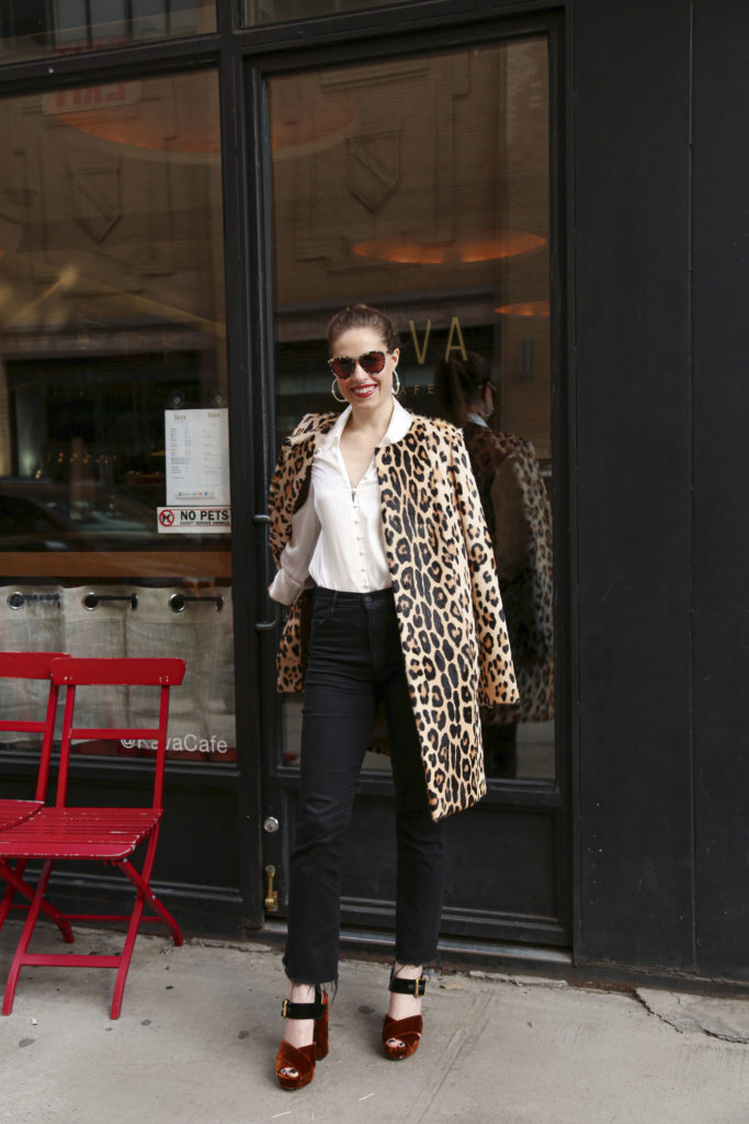 Woman standing outside a coffee shop wearing a leopard coat with a white silk top, black jeans and black & orange velvet shoes