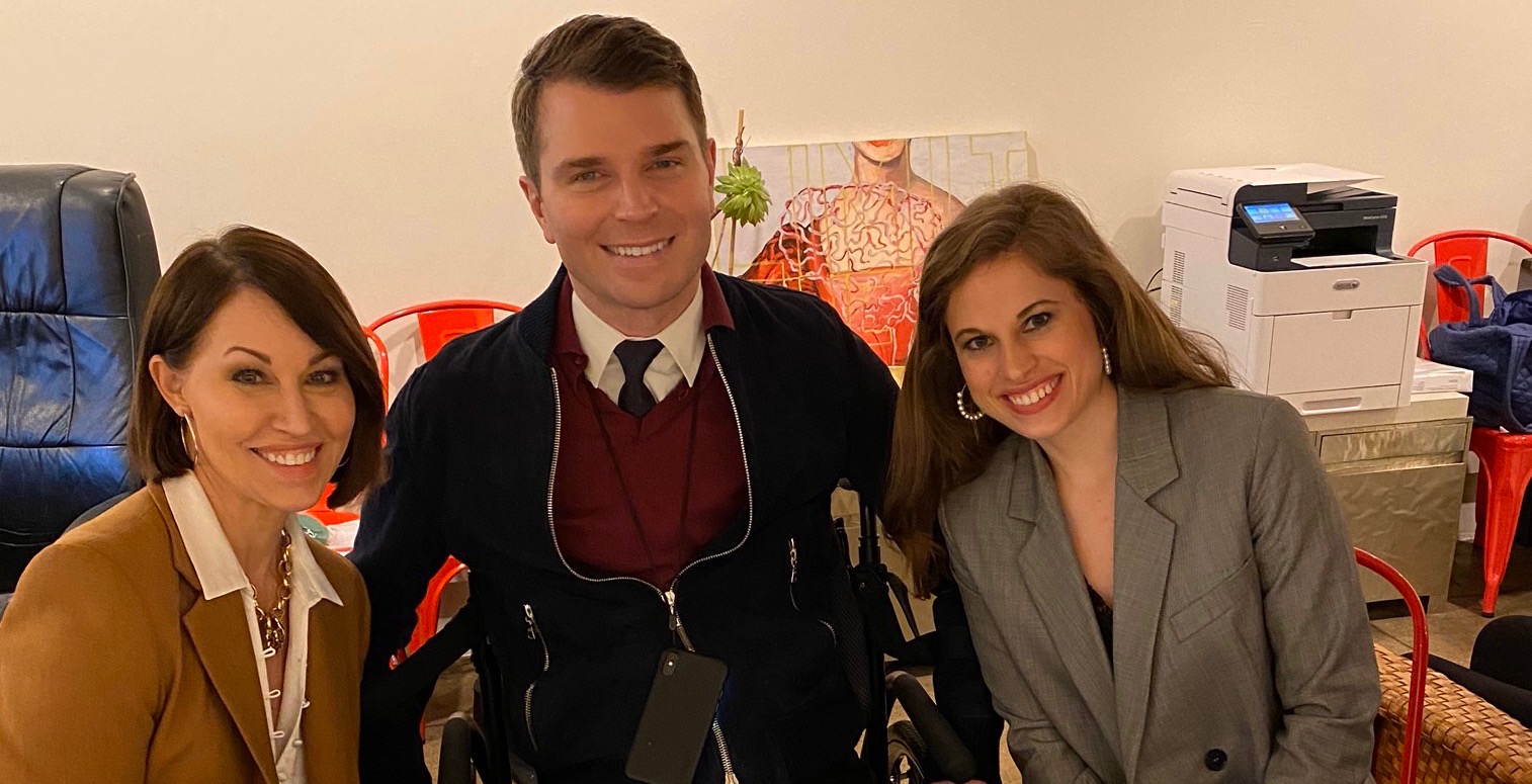 Photo of three people: A women in a camel colored blazer, a man in a wheelchair & a woman in a grey blazer.