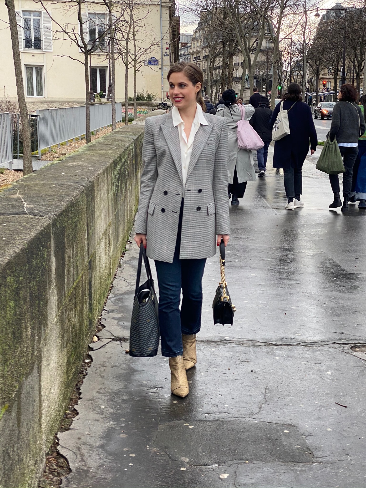 Woman walking down the street in Paris wearing a plaid blazer with a white-button down underneath with jeans and black & gold sparkle booties. Holding a purse in each hand