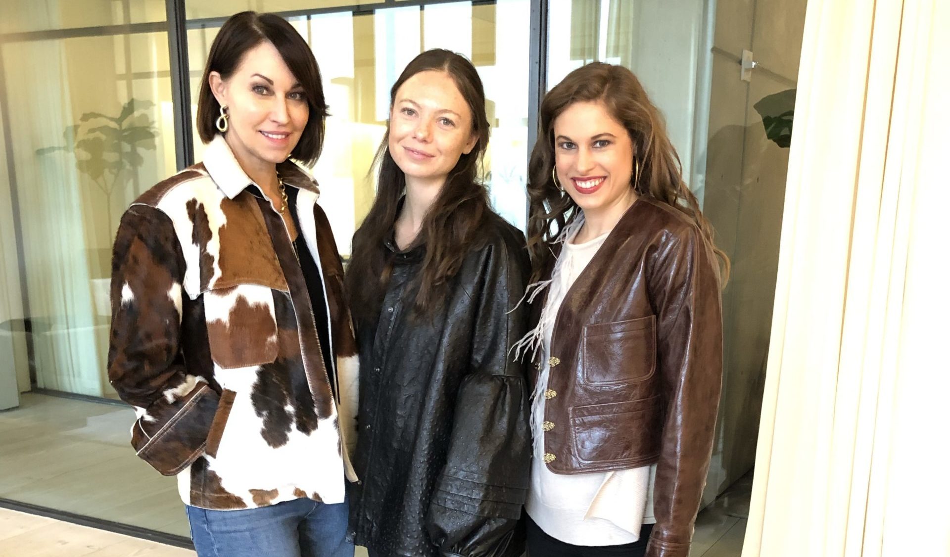Picture of three women. One wears a cowhide pattern jacket, another wears a black leather jacket and the last with curly hair wears a brown cropped leather jacket