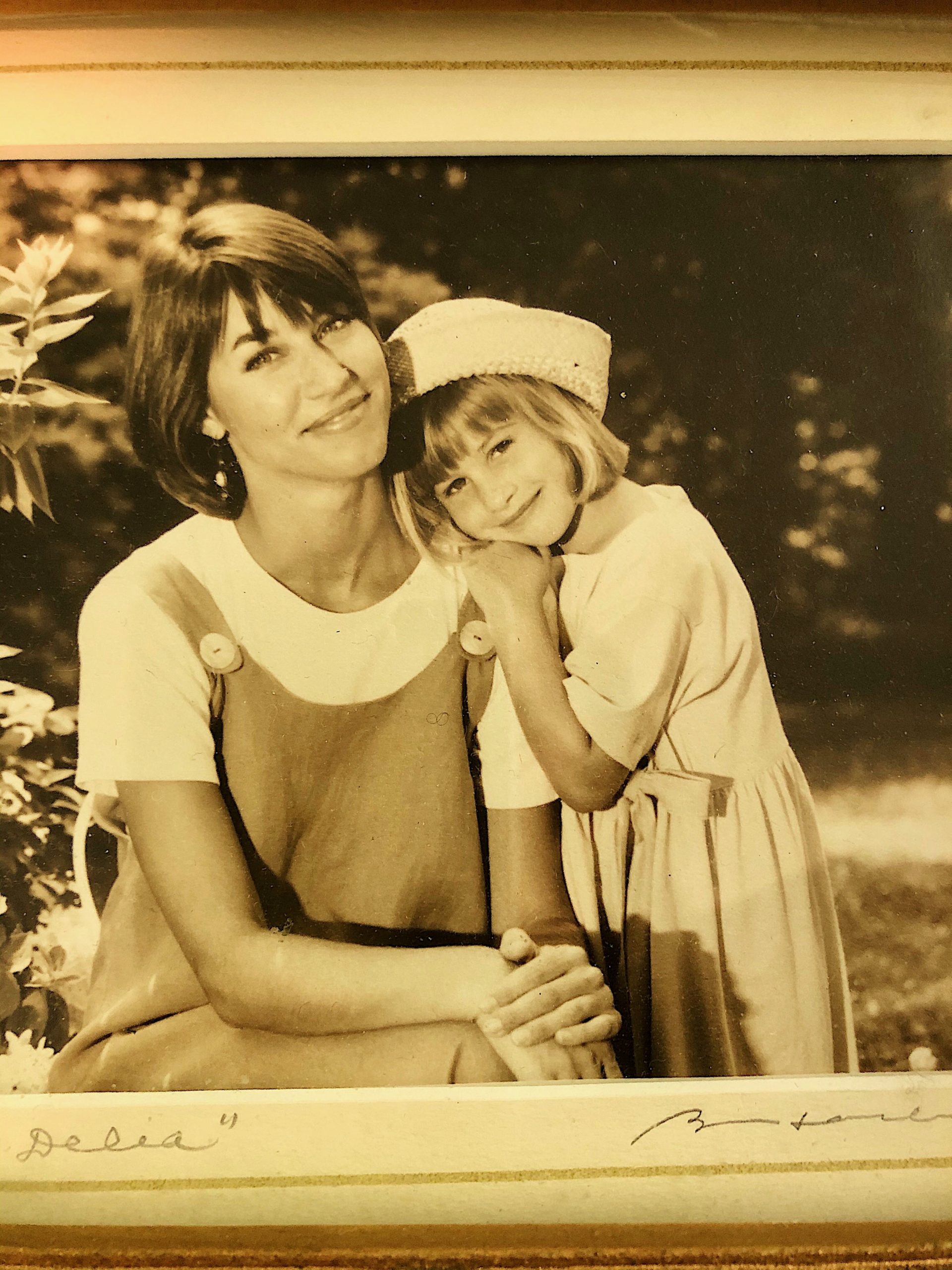 Mother & daughter: both with brown hair - one wears a dress over a short-sleeved shirt and the daughter wears a hat & a dress. The daughter has her mother's head on her shoulder