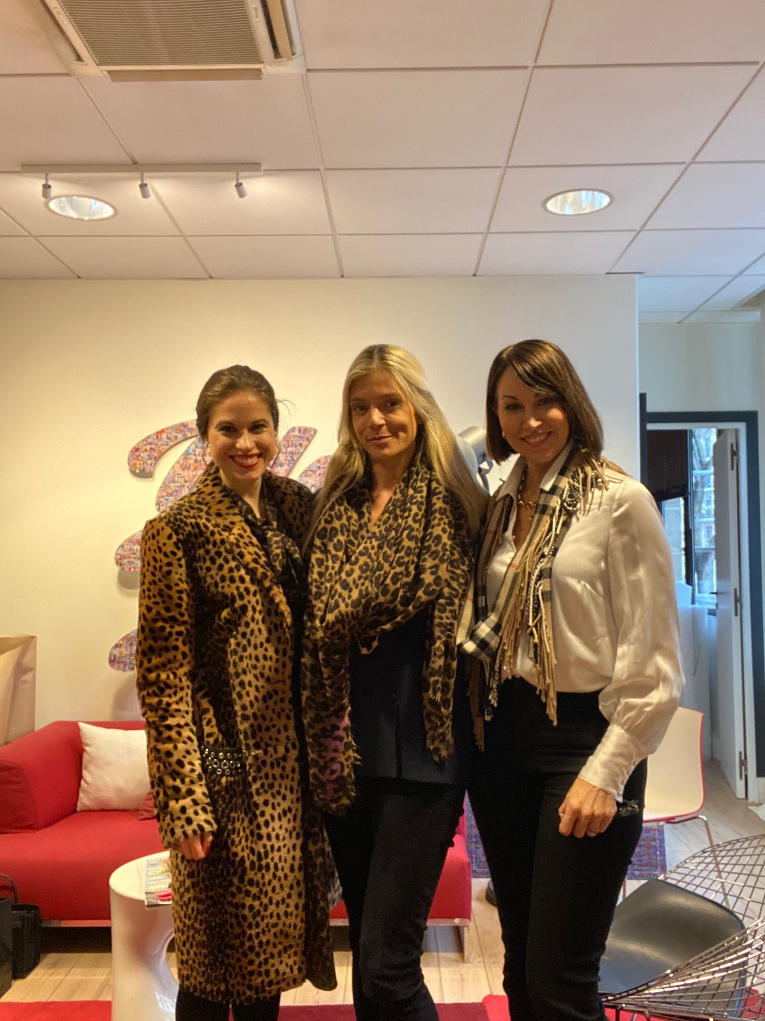 Three women standing together smiling: two with brown hair & one with blonde. One wears a leopard print coat, another wears a leopard print scarf & jeans and the last wears a burberry scarf with an ivory silk blouse