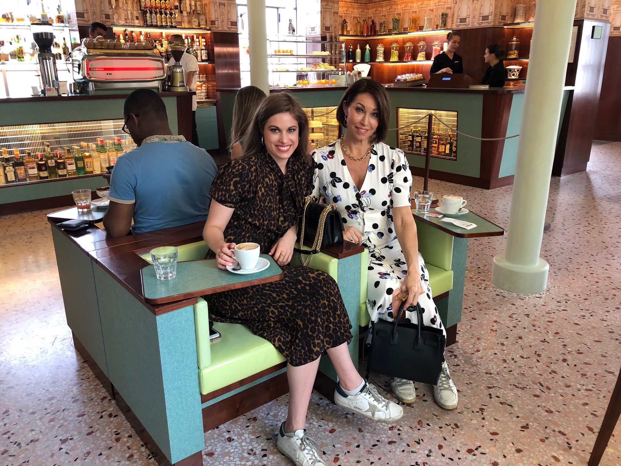 Two women sitting at a restaurant drinking coffee: both with brown hair. One wears a leopard print short-sleeve dress, the other wears a black & white printed short-sleeve dress, both wearing sneakers