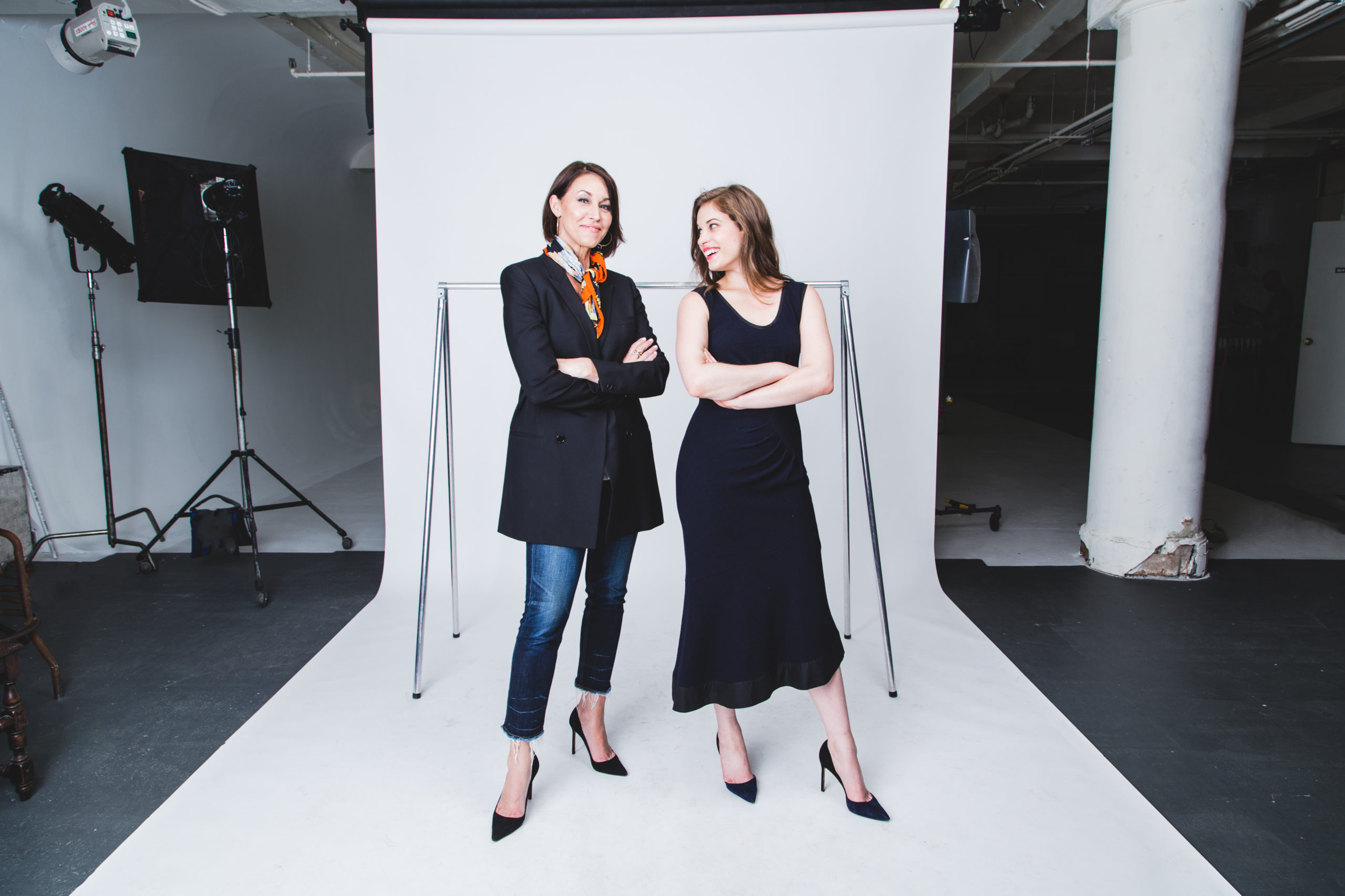 Two women standing on set in front of a clothing rack: one wearing a black blazer, jeans and black pumps; the other wearing a navy dress with navy pumps. Both have brown hair
