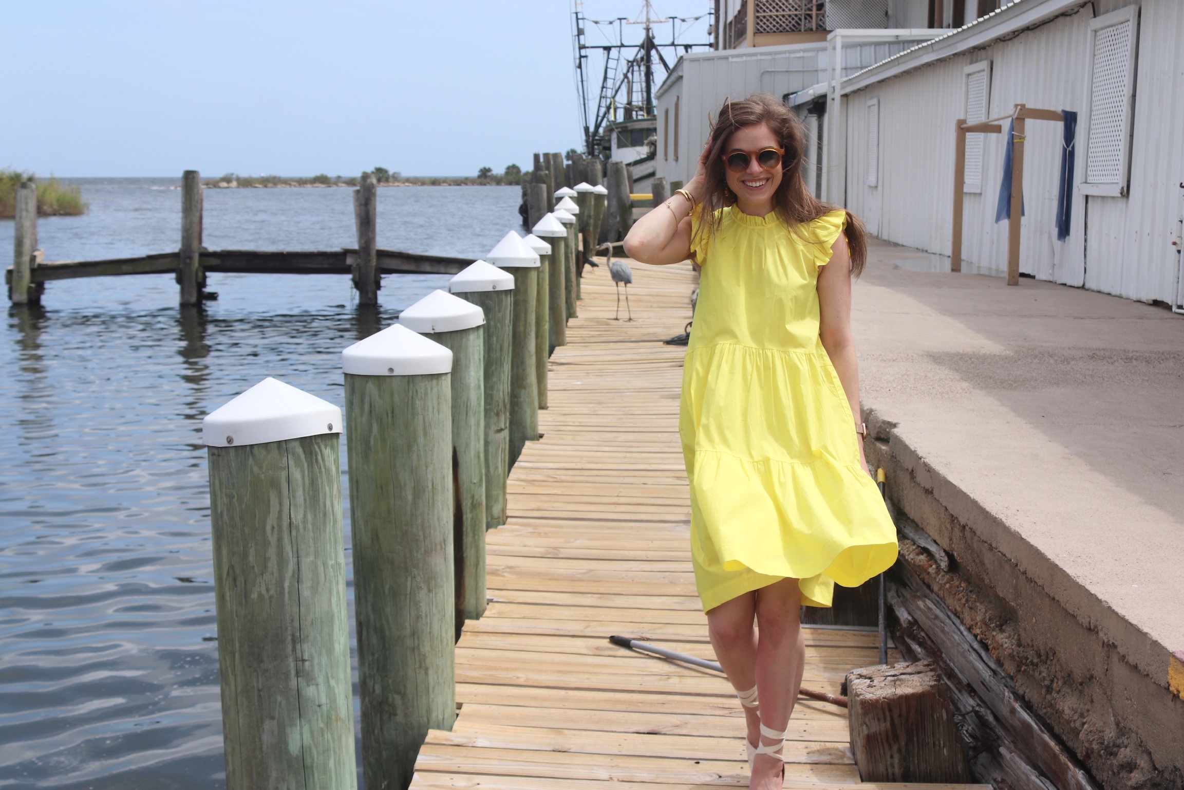 Girl walking down a dock wearing a pink dress & espadrilles with brown hair. A blue heron is behind her