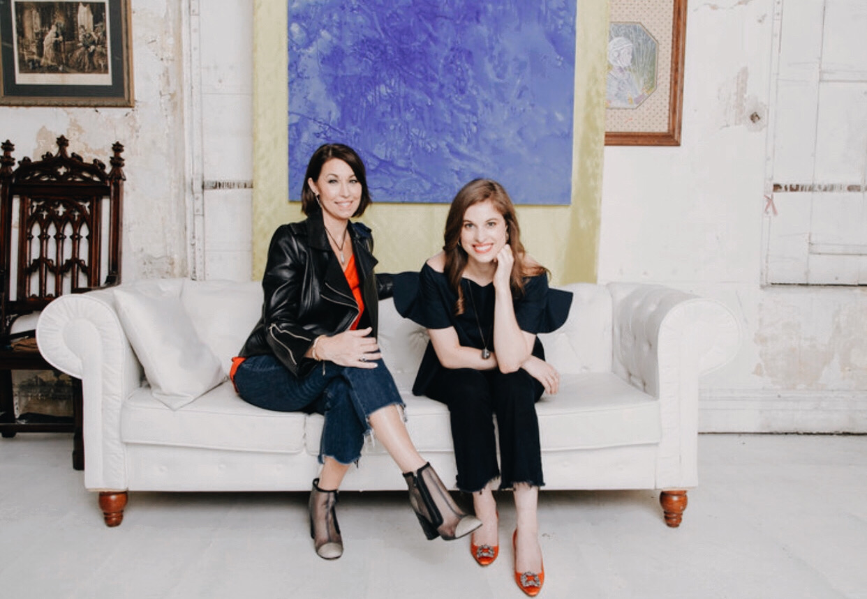 Two women sitting on a couch: they both have brown hair. One wears a black leather jacket with an orange top and booties. The other wears a navy top with jeans and orange pumps