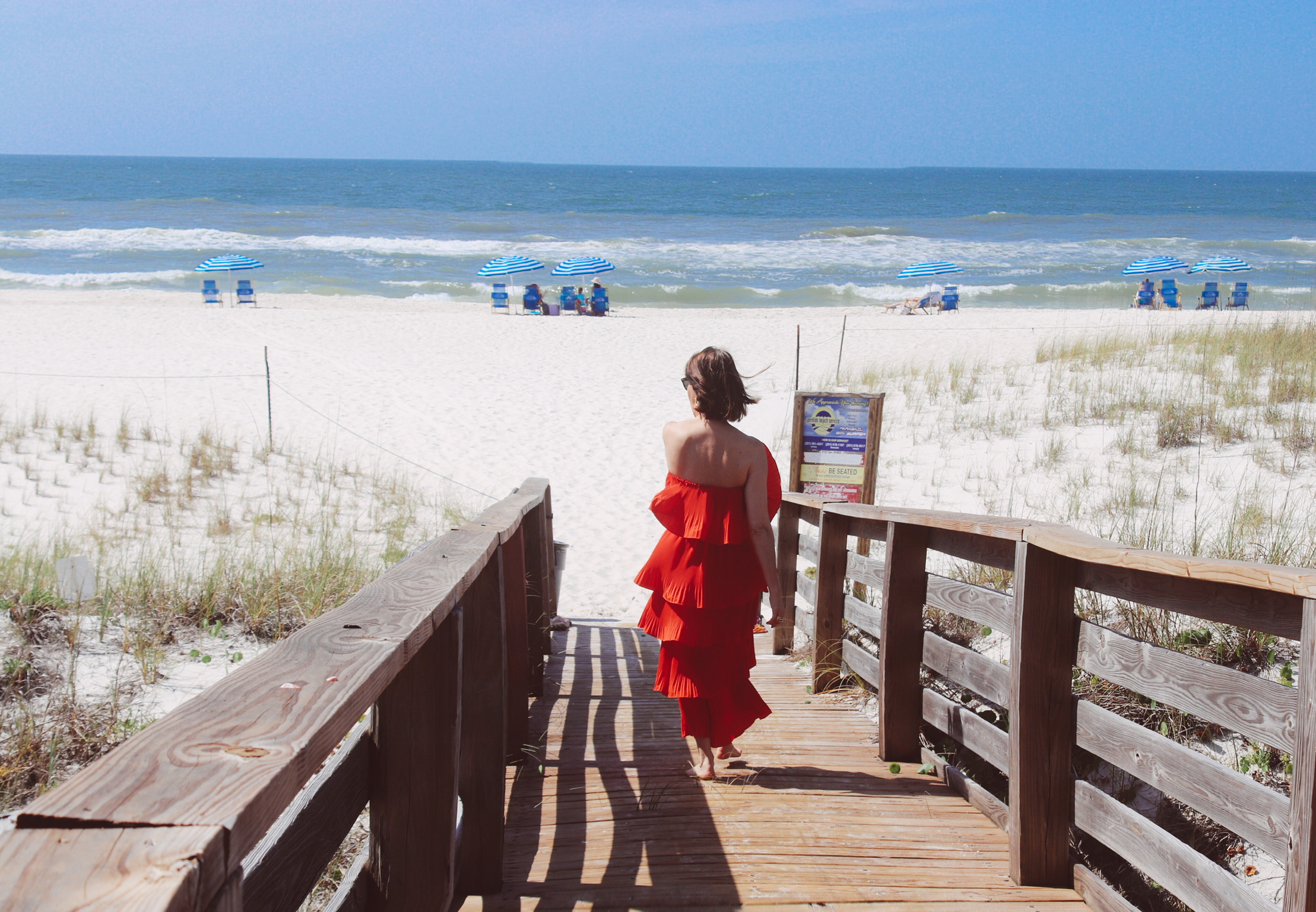 Woman with brown hair wearing a red tiered dress walking on the boardwalk down to the beach