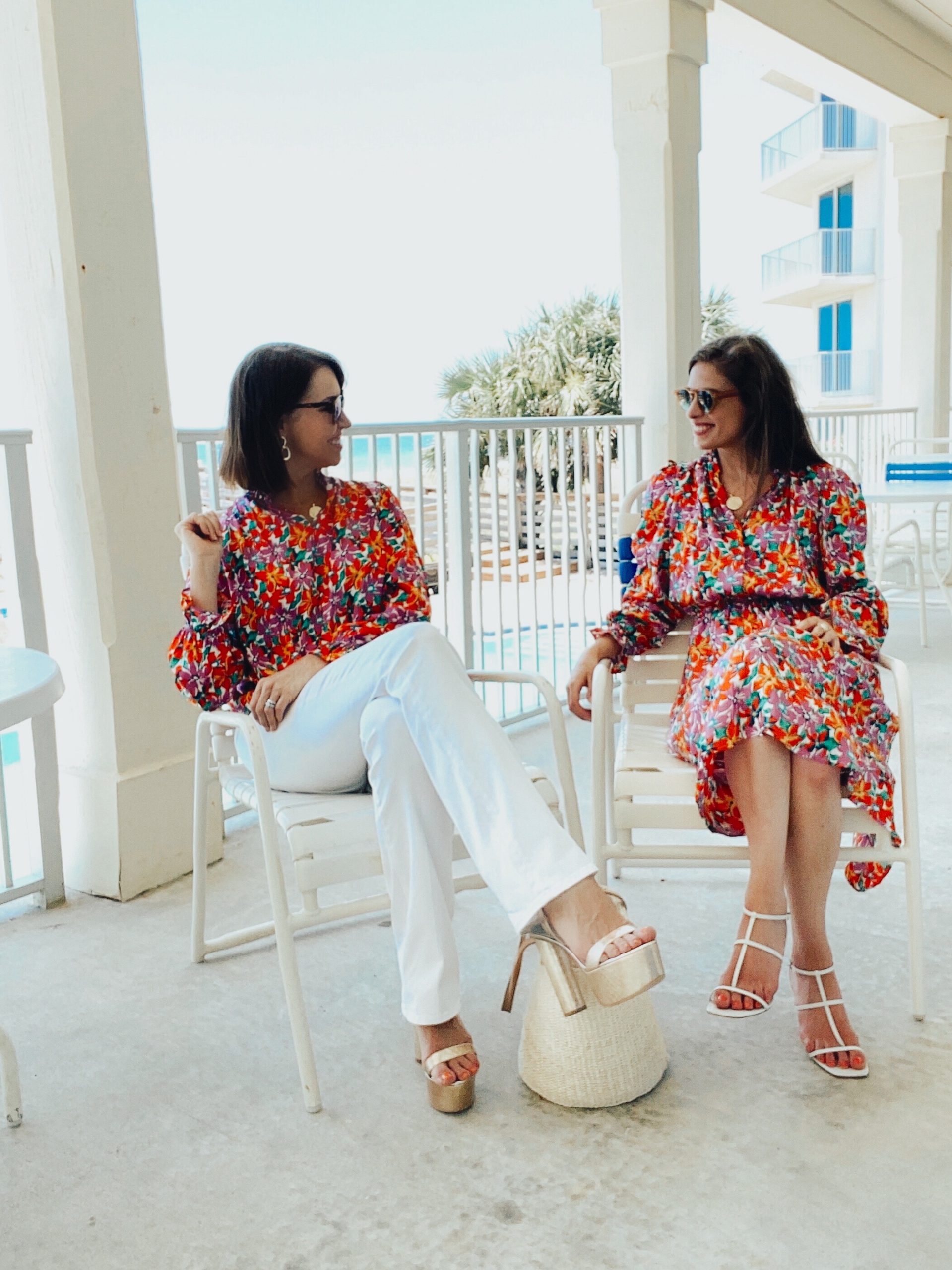 Two women with brown hair wearing bright florals sitting on a deck over looking a pool