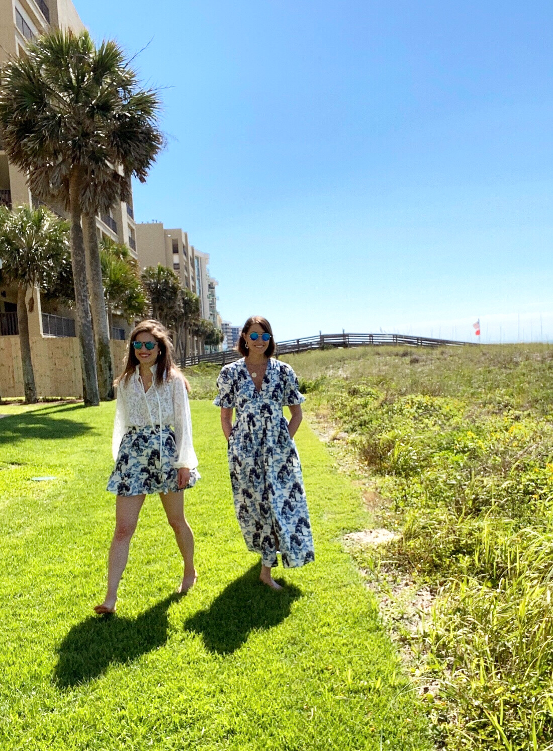 two women walking in the grass at the beach, both have brown hair and are wearing sunglasses. One wears a white top tucked into a short blue printed skirt and the other wears a blue and white printed dress. both are barefoot