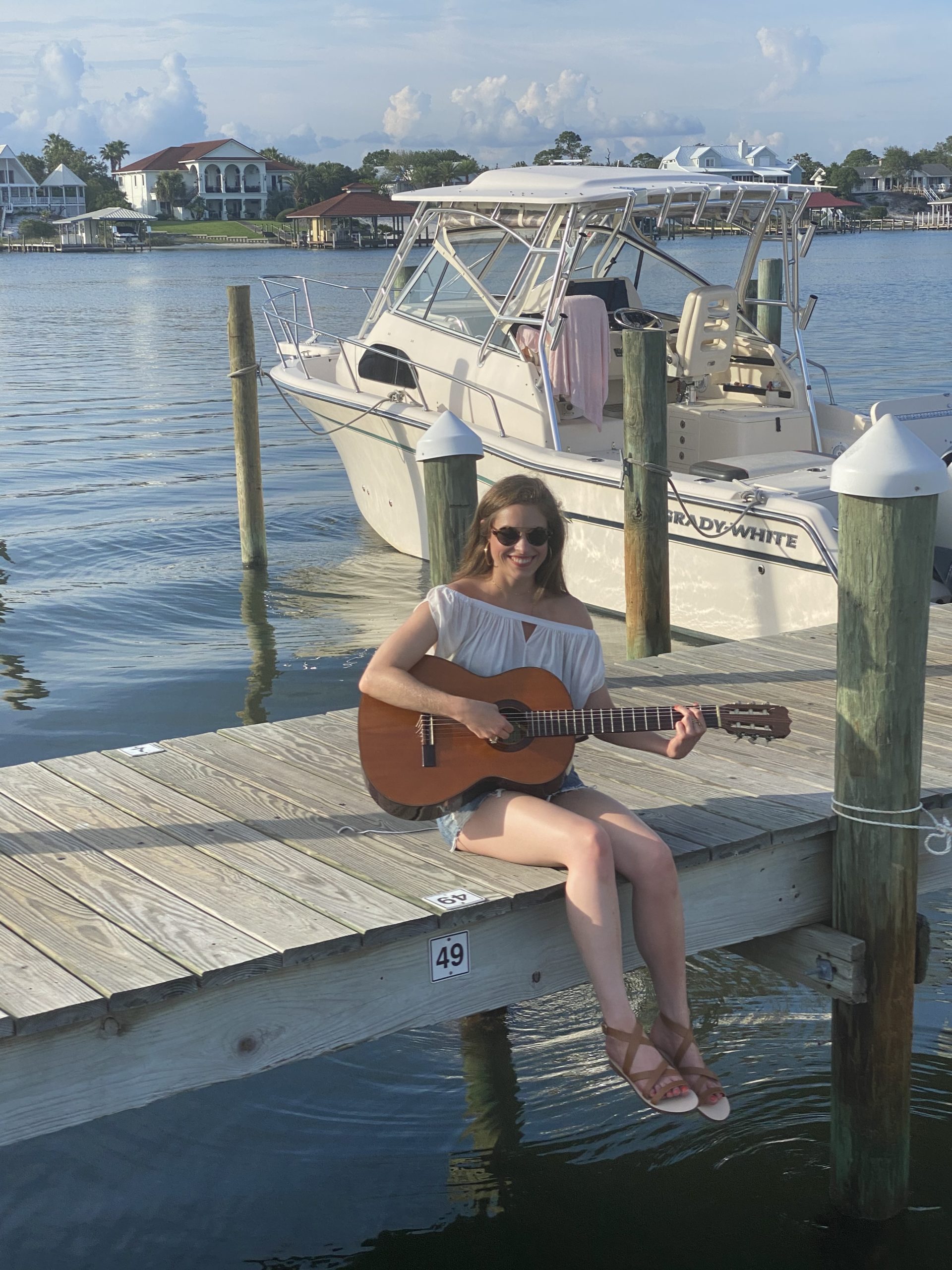 Girl with brown hair sitting on the dock of the bay wearing a white off the shoulder top, blue jean skirt, brown sandals playing acoustic guitar