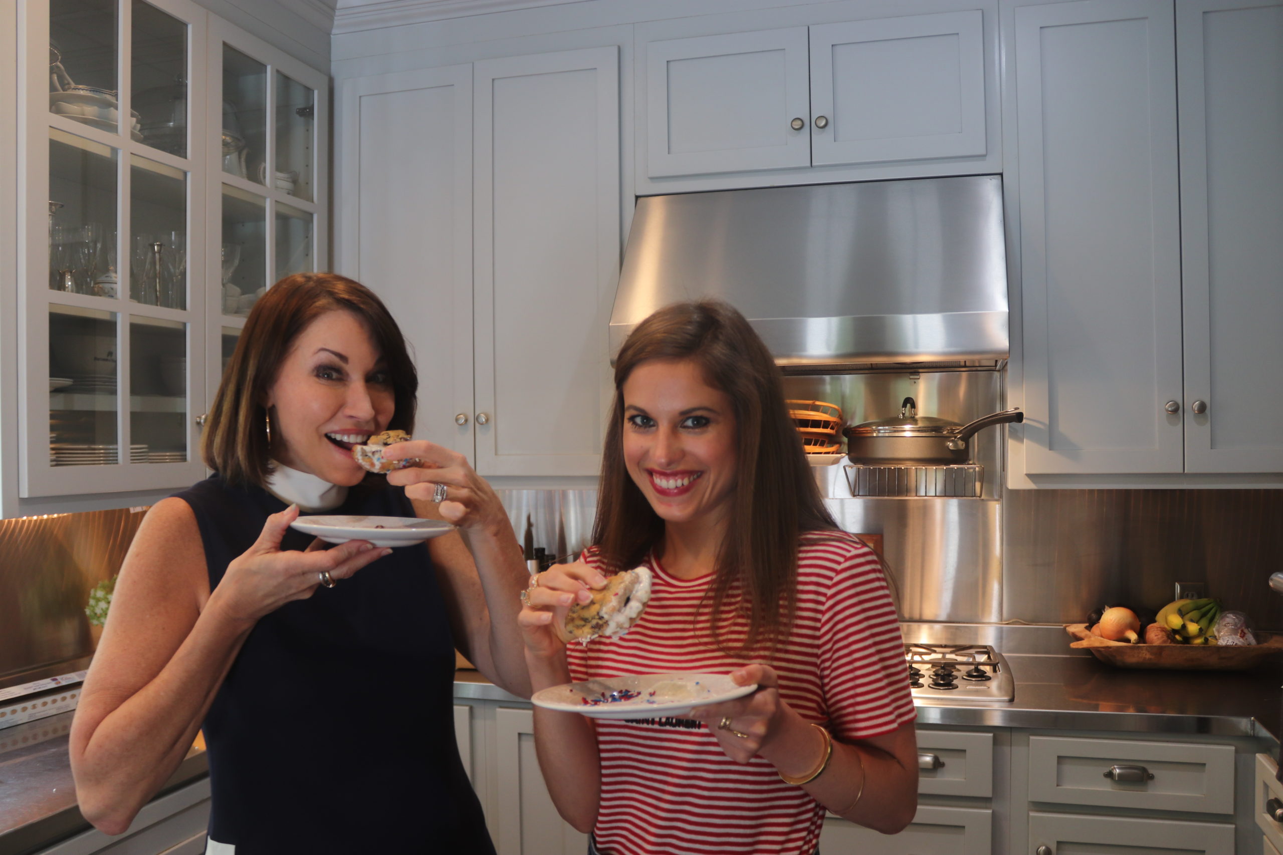 Two women standing in a kitchen, both with brown hair. One is wearing a navy top with a white collar and the other is wearing a red and white striped shirt. They are eating 4th of july themed ice cream sandwiches