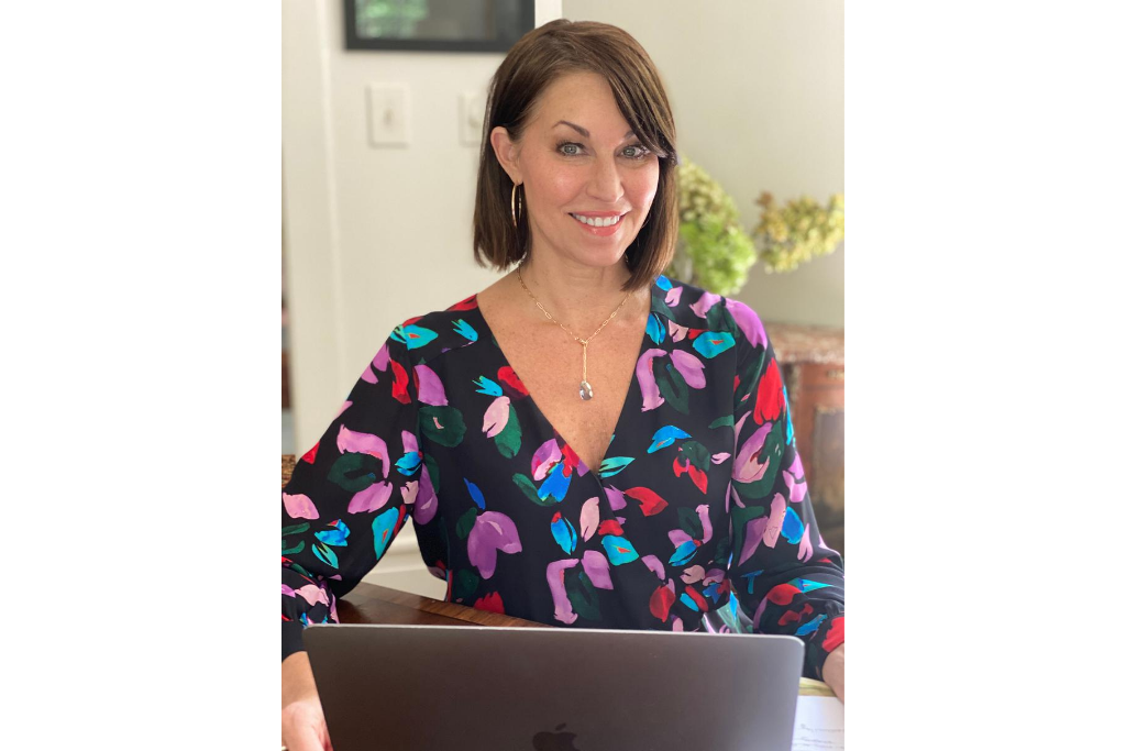 Woman with brown hair smiling and sitting working at her computer wearing a lariat necklace and colorful printed top