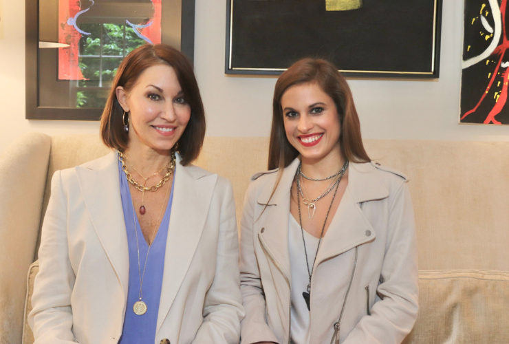 Two women both with brown hair sitting on a couch wearing layered necklaces: one wears a white blazer with a blue shirt and the other wears a cream colored leather jacket