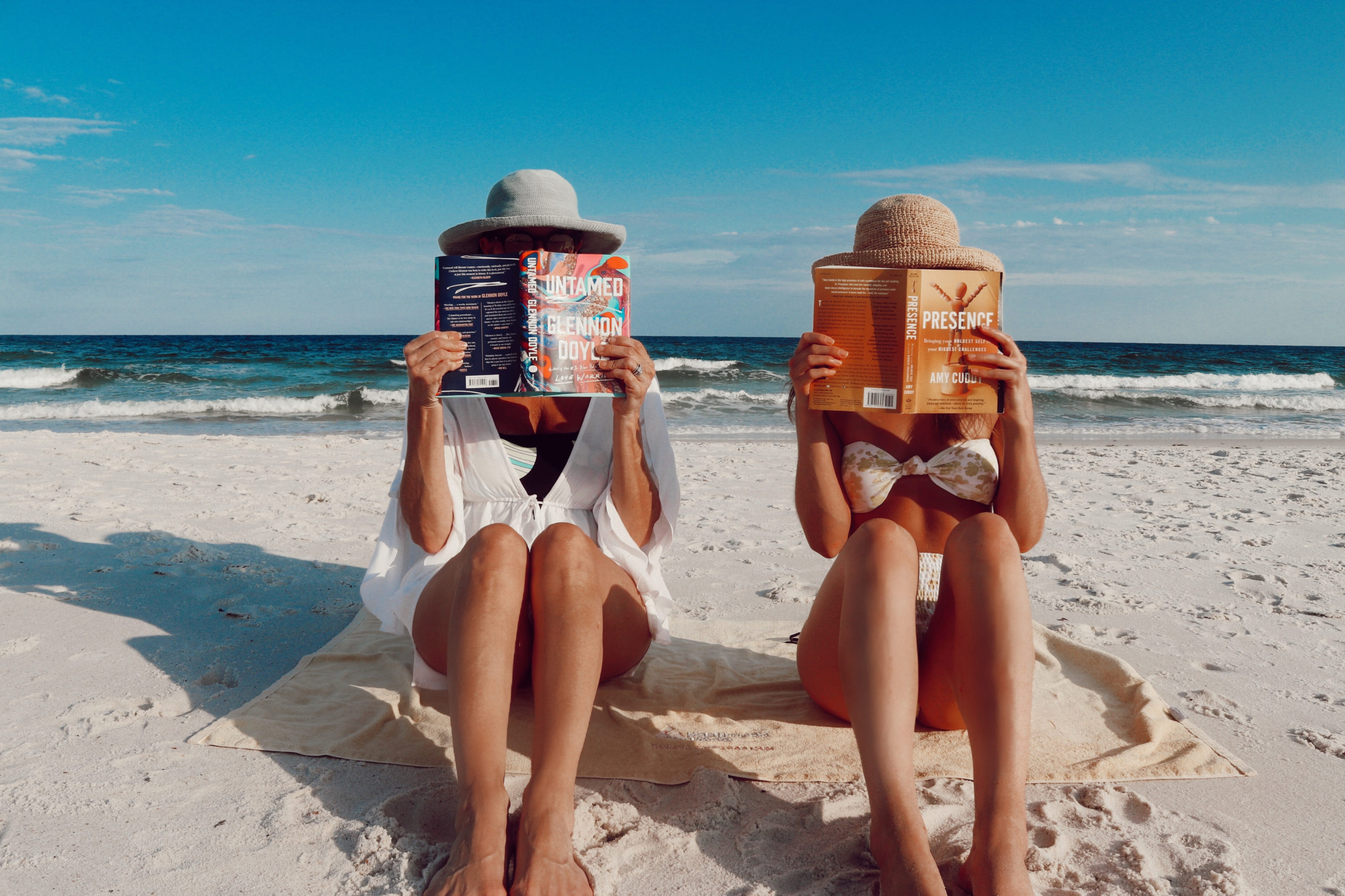 Two women sitting on the beach wearing bathing suits and sun hats holding up books
