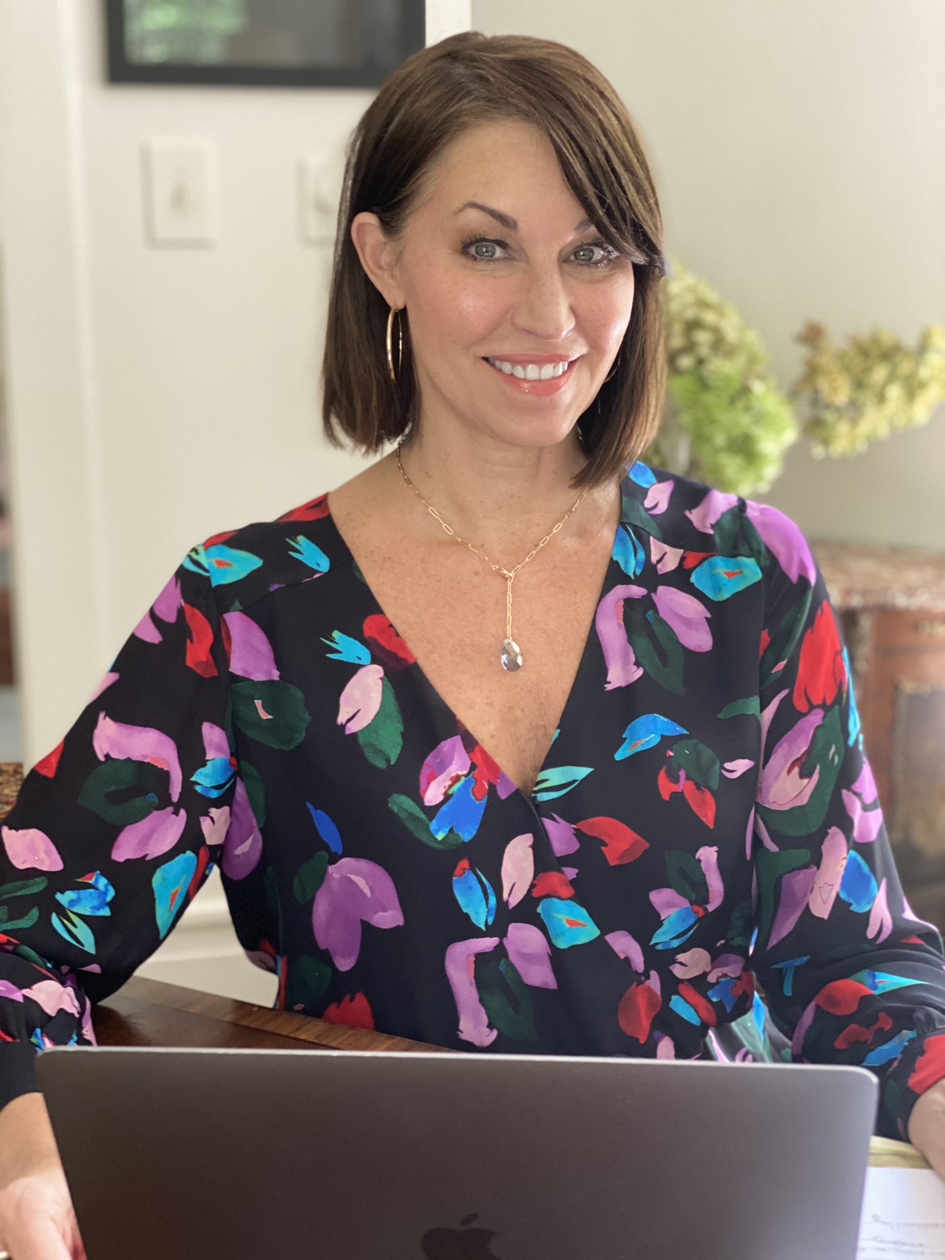 what to wear on zoom Woman with brown hair smiling and sitting working at her computer wearing a lariat necklace and colorful printed top