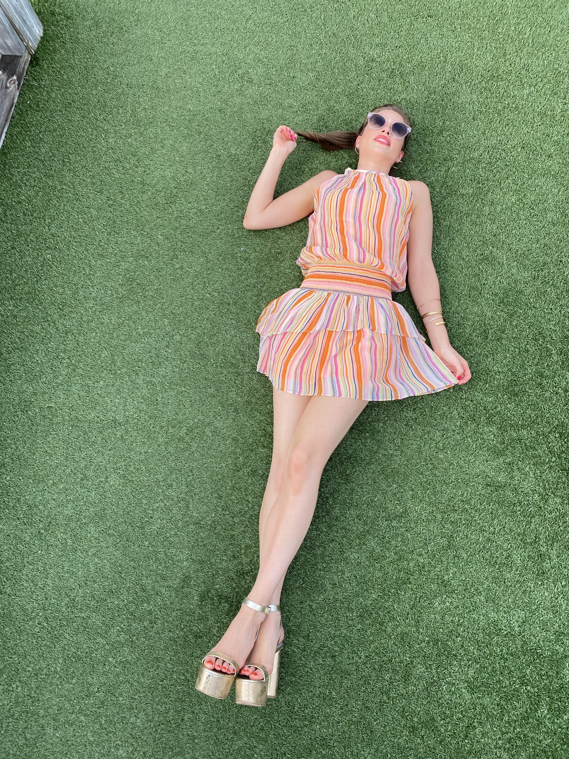 Woman lying on the ground wearing a sleeveless colorful striped dress with pink sunglasses, a ponytail with brown hair and wearing gold platform shoes