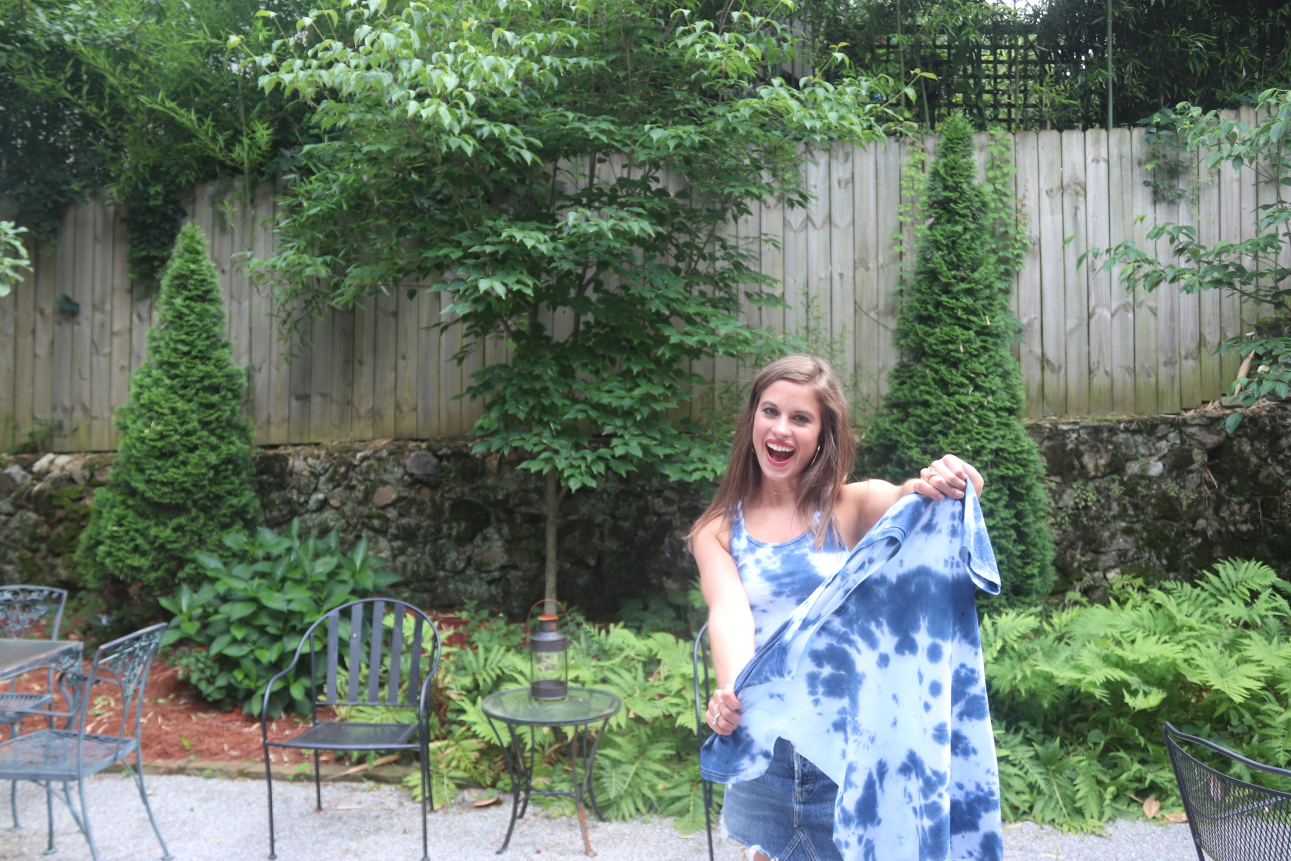 Girl standing outside with brown hair wearing a tie-dye tank & holding a tie dye t-shirt