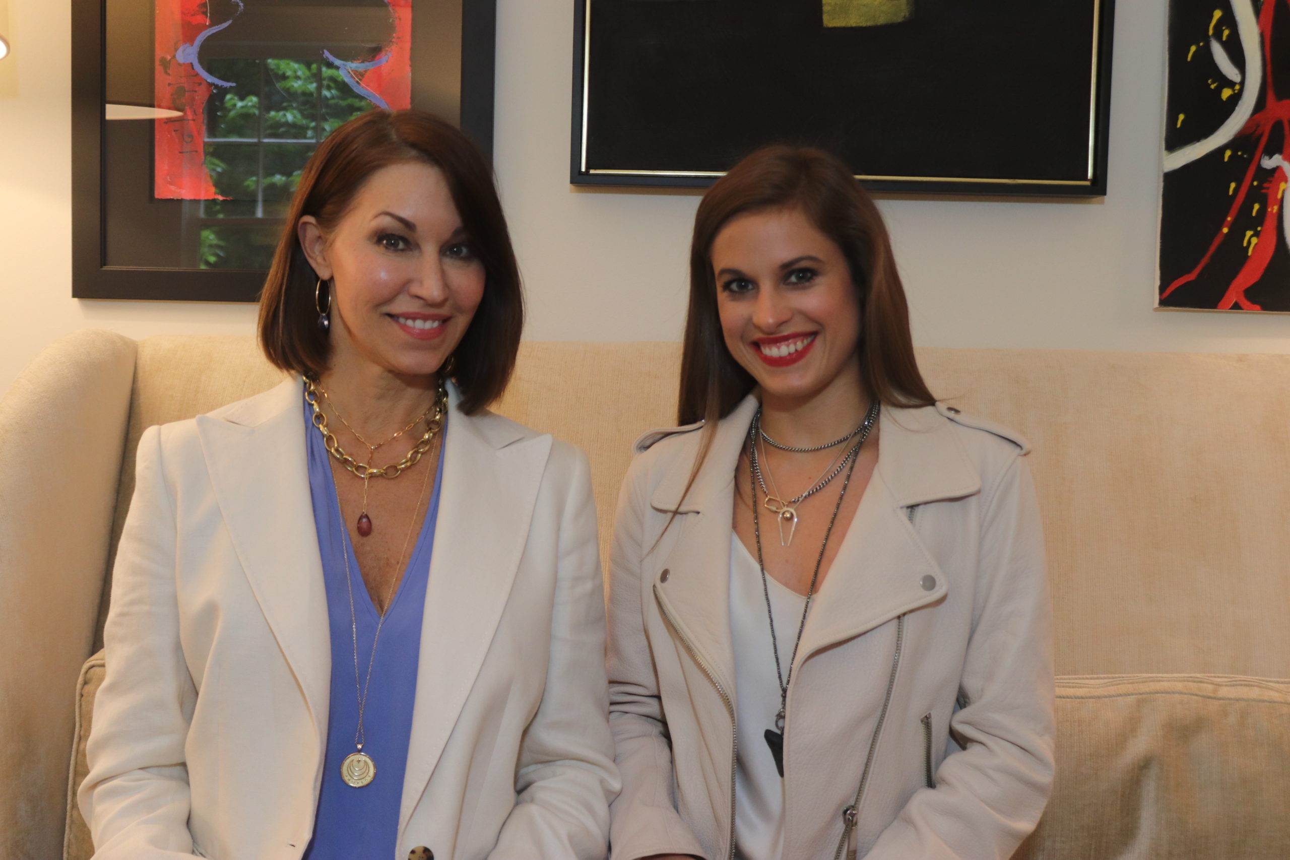 Two women both with brown hair sitting on a couch wearing layered necklaces: one wears a white blazer with a blue shirt and the other wears a cream colored leather jacket