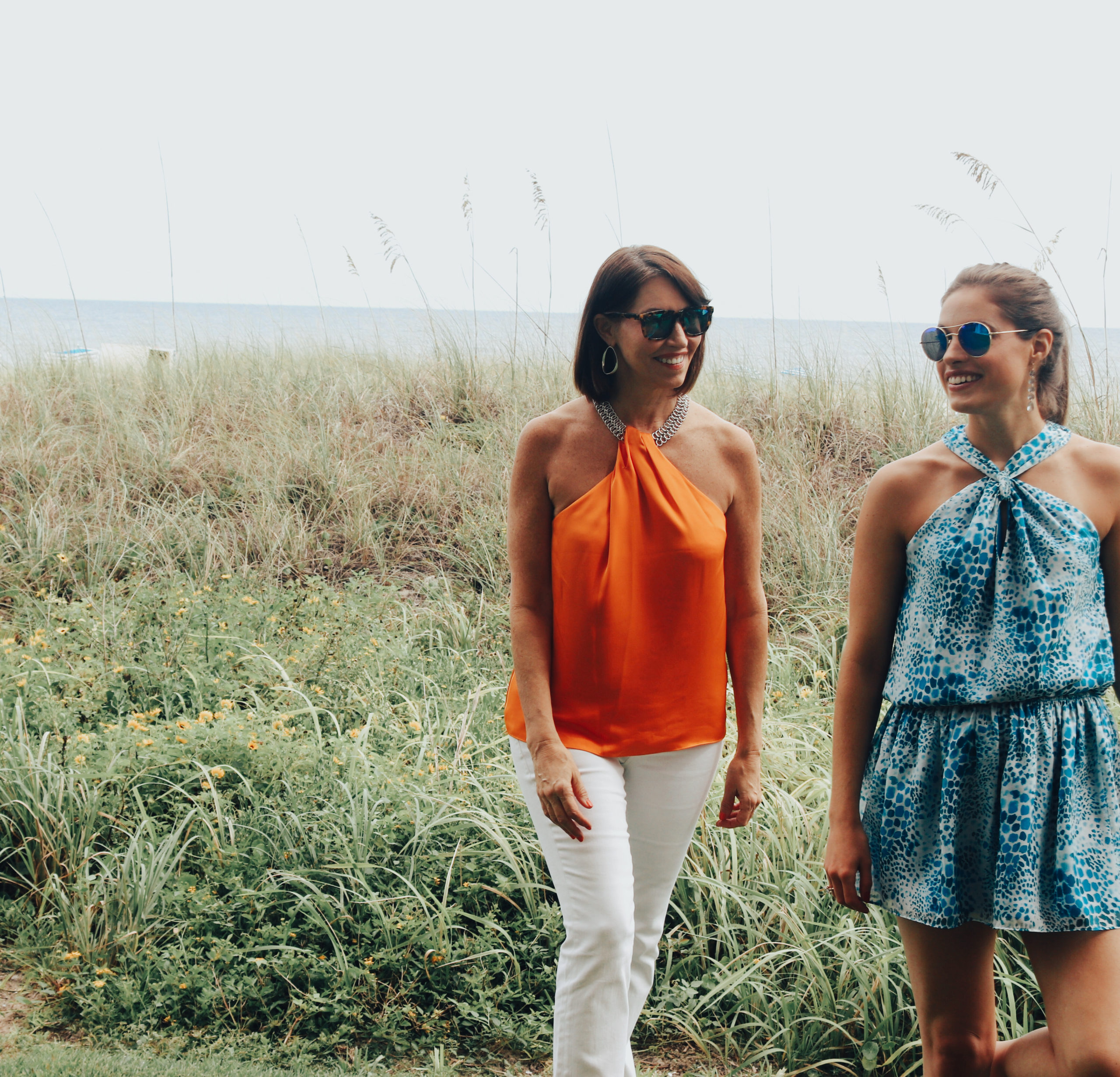 Two women walking in the grass: one wears an orange top with white jeans & the other wears a blue dress. They both have brown hair & are wearing sunglasses