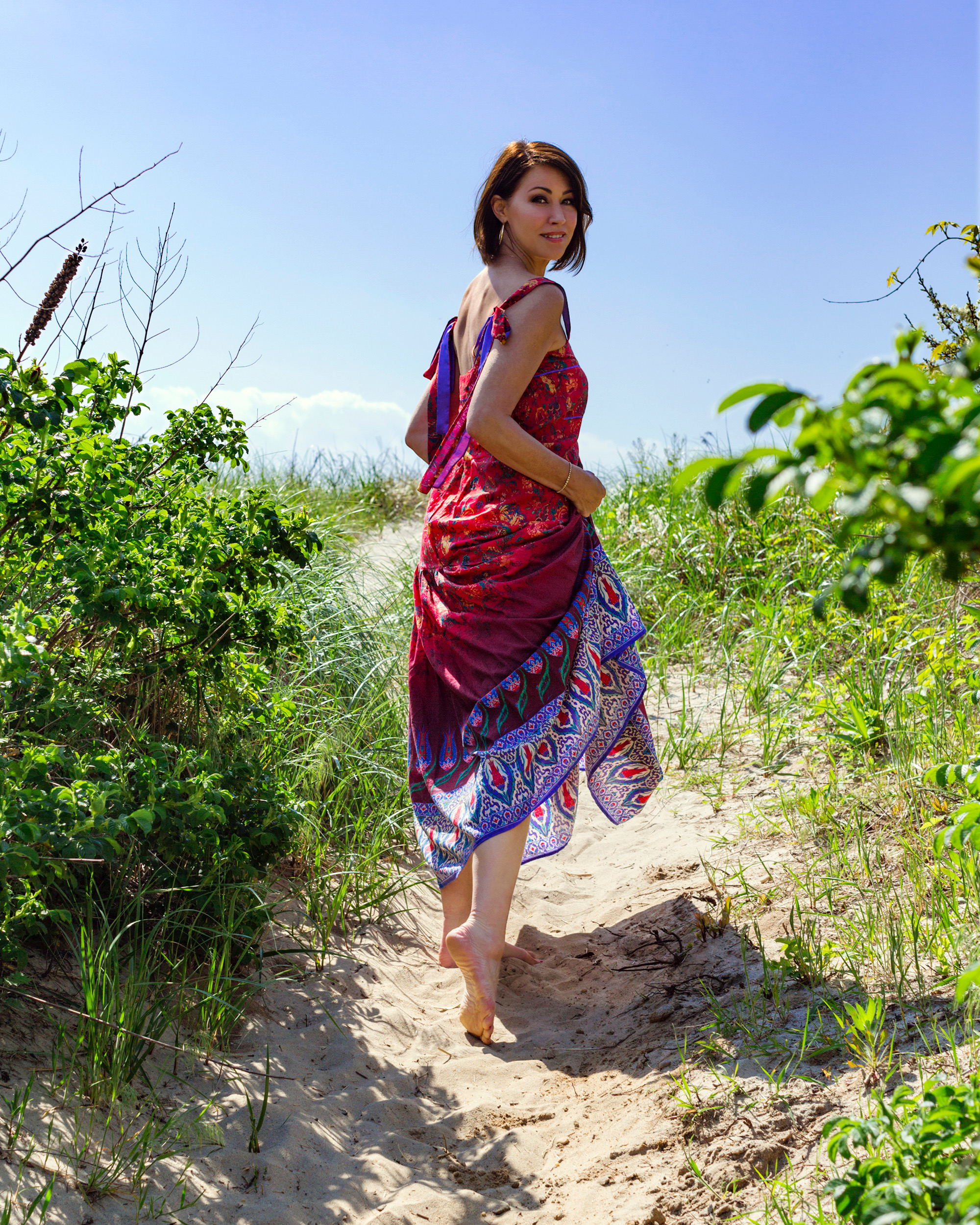 Woman with brown hair wearing a red printed sleeveless dress walking at the beach