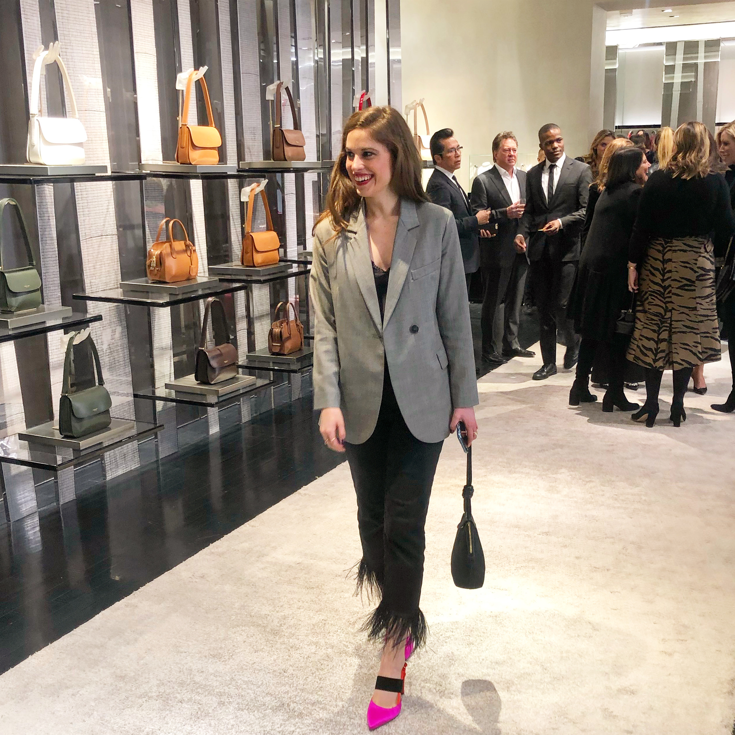 Woman walking through a store wearing a grey blazer, jeans with feathers, a black purse and pink shoes