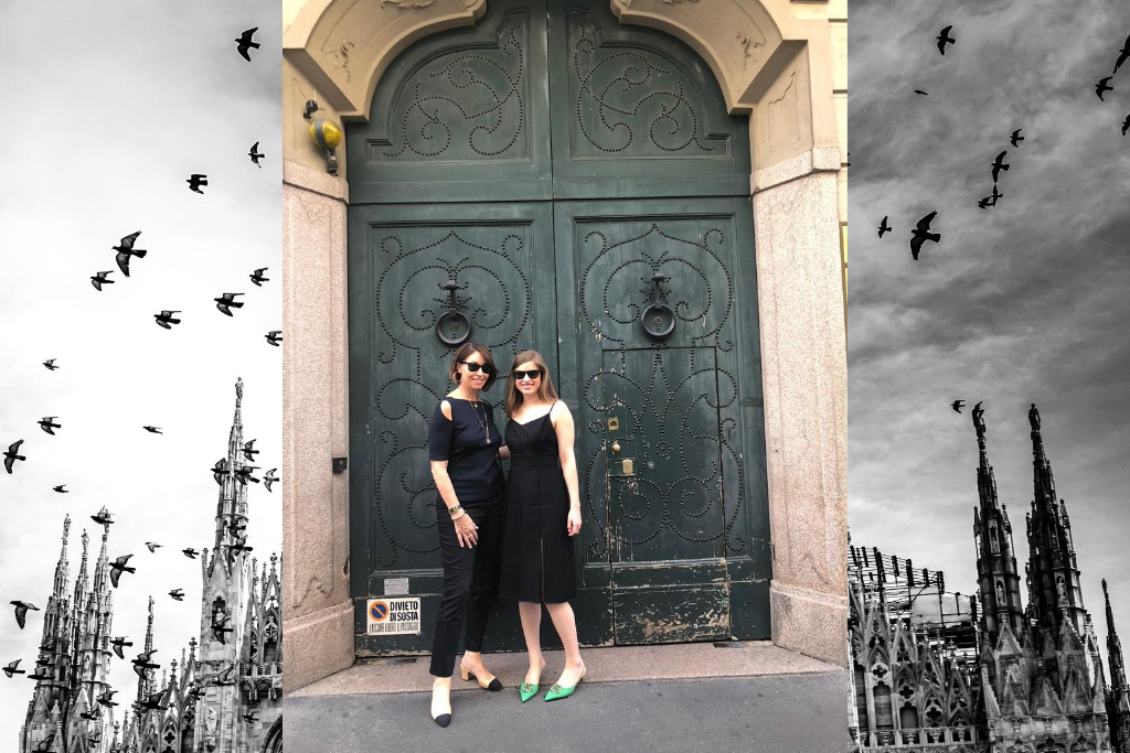 Two women wearing black sunglasses in front of a dark green door. One girl wears a black dress with green shoes & the other wears a navy top with cutouts, jeans & slingback shoes