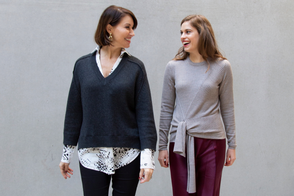 Two women looking at each other and walking together, both have brown hair. One wears a grey v-neck sweater and the other wears a beige sweater and wine colored skirt