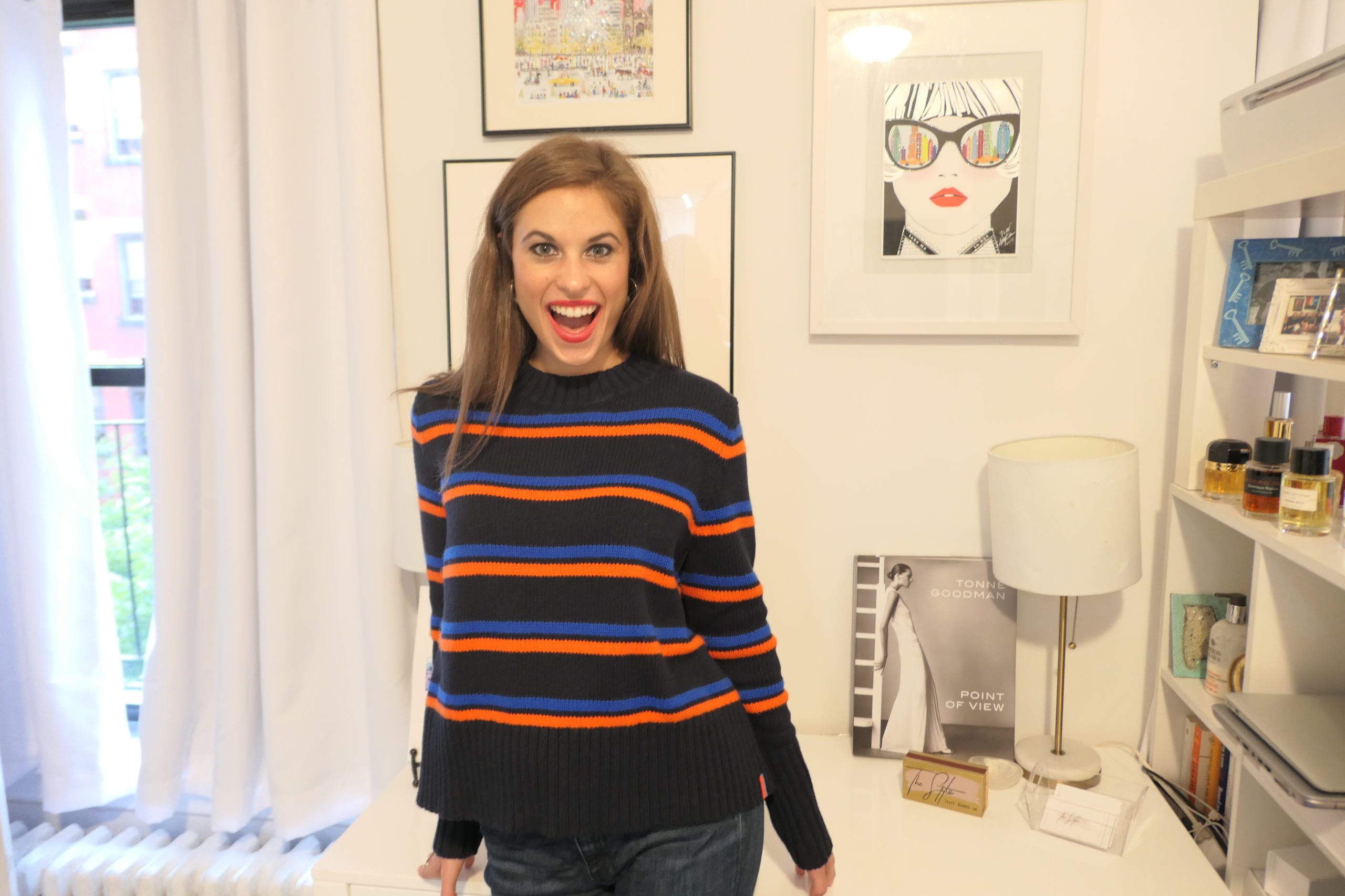 Woman standing at a desk with art hung on the walls with brown hair wearing a black, blue & red striped sweater and jeans. She is smiling