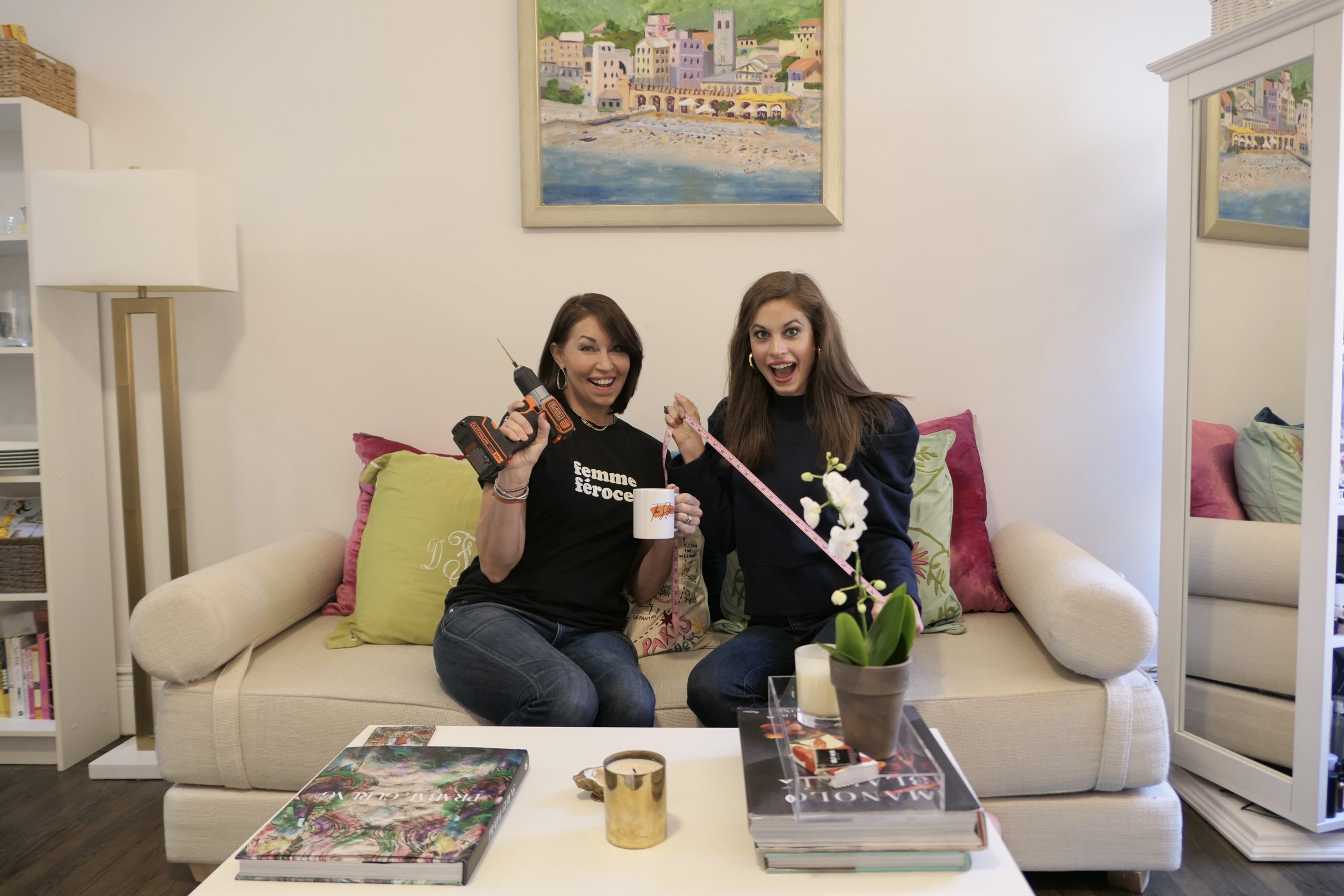 Two women both with brown hair sitting on a couch holding tools, one wears a black tee and jeans, the other wears a sweatshirt & jeans.