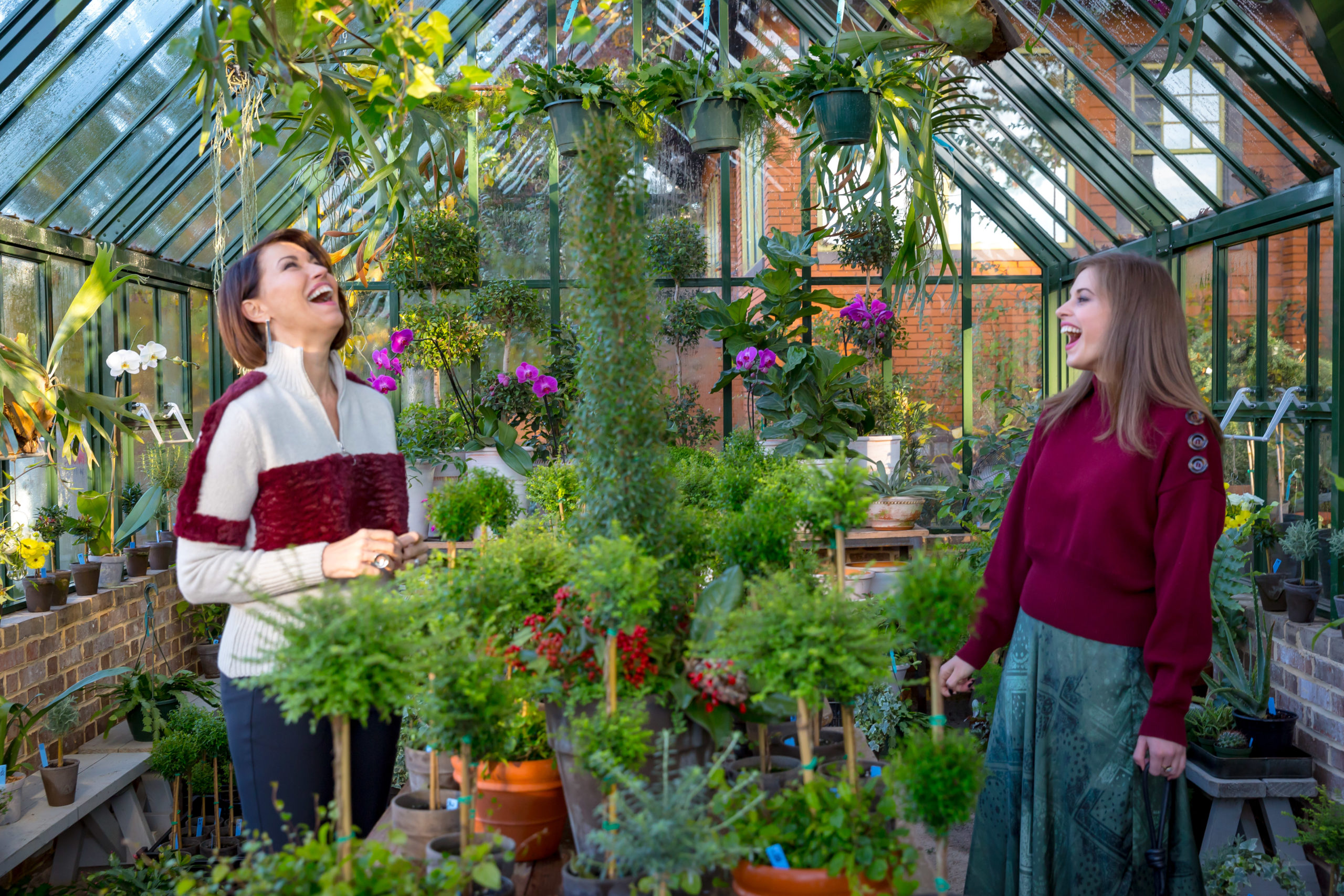 Two women in a greenhouse both with brown hair laughing. One wears a wine & grey sweater and the other wears a wine sweater and a green skirt