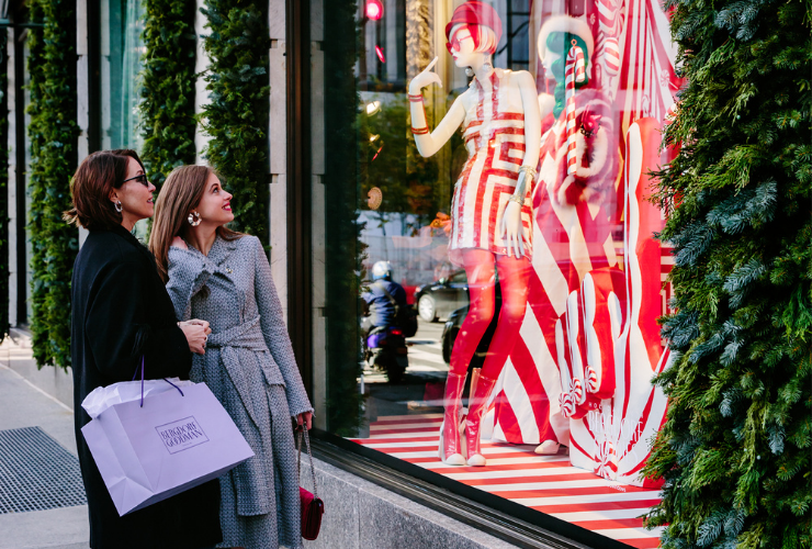 Two women walking and looking at the Bergdorf Goodman windows both have brown hair and wearing coats and heels