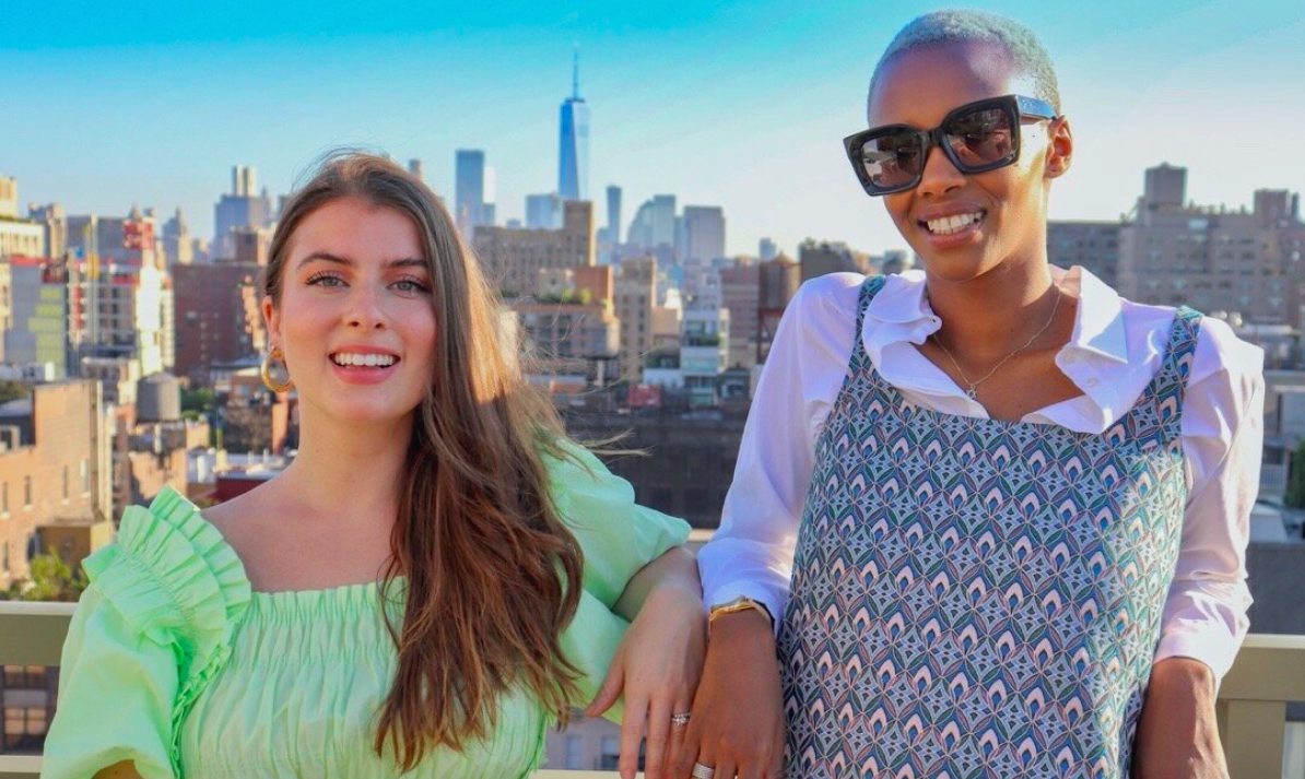 Two women standing on a rooftop in NYC: one white woman with brown hair and one black woman wearing sunglasses.