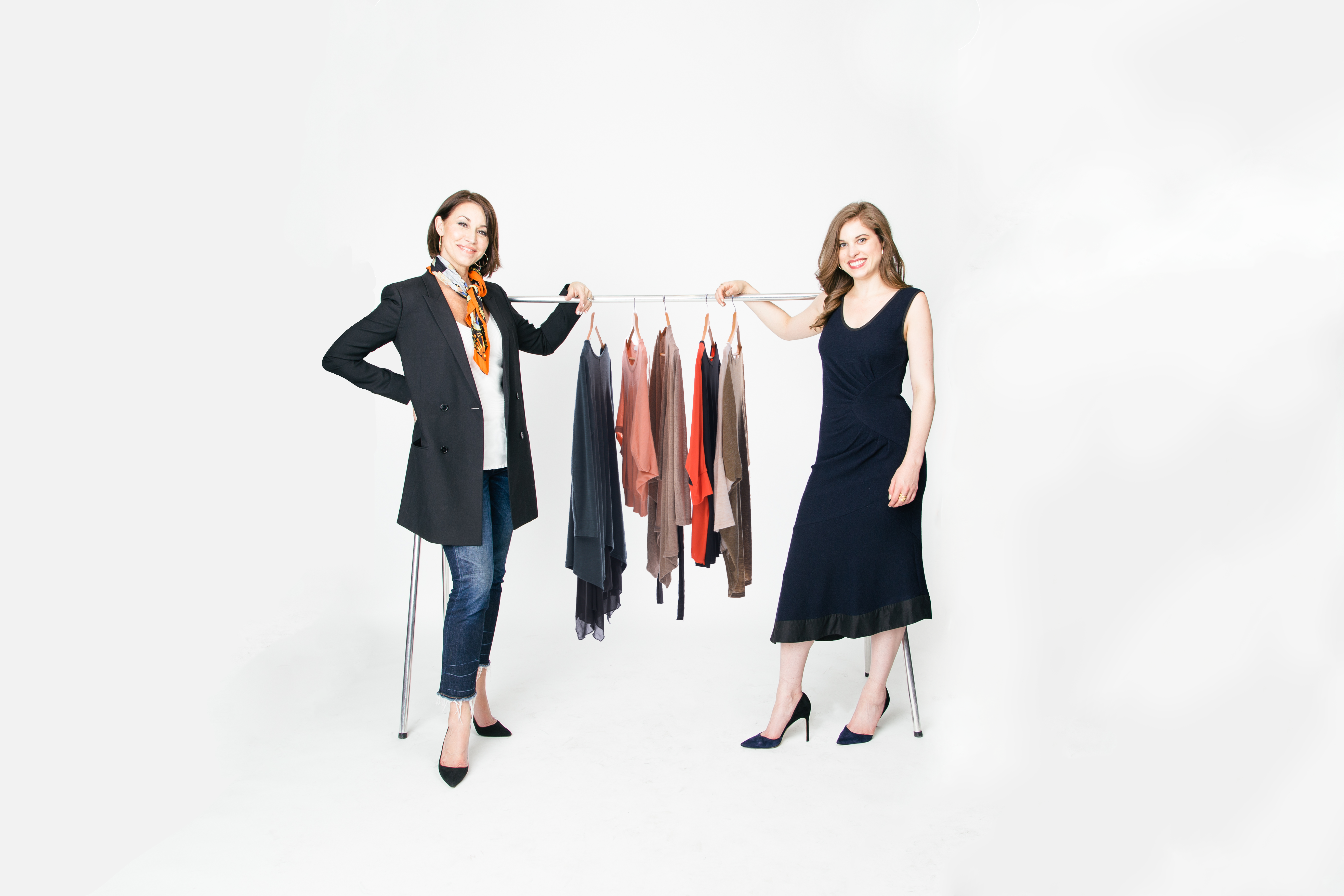 Two white women with brown hair standing in front of a rack of clothing. One wears a navy dress with heels and the other wears a black blazer, white camisole, jeans and heels