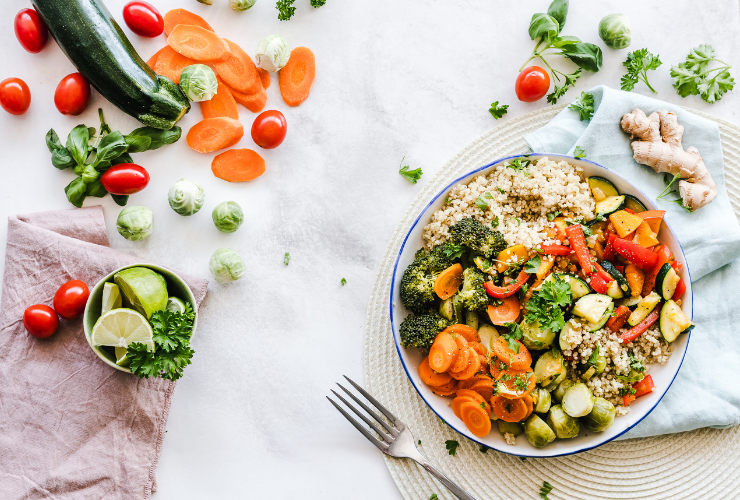 Bowl of a salad filled with quinoa and fresh vegetables on a white placemat with a fork next to it, there are also fresh veggies laying on the table