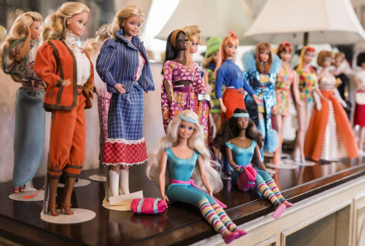 A bunch of Barbie dolls sitting on a mantle. They are made up of all different ethnicities and are wearing a variety of outfits.