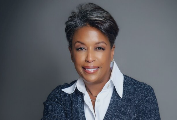 Black woman with short hair (grey/black) looking at the camera and smiling, wearing a grey sweater over a white button-down collared shirt