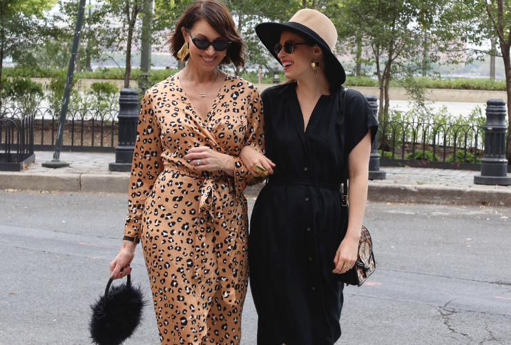 Two women walking down the street in NYC. They are white and have brown hair. One wears mini black sunglasses and a v-neck long-sleeve beige and brown leopard print dress. She wears black pumps and a black round feather bag. The other girl wears a black and camel hat with a black short-sleeve v-neck dress with cowboy boots. They are locking arms and smiling