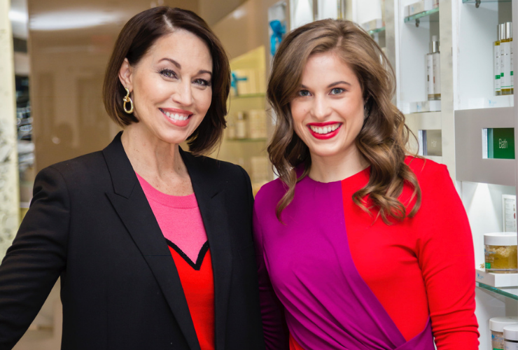 Two white women smiling: both with brown hair. They are in a store and one wears a black blazer with a red and pink heart sweater. The other wears a pink and red dress