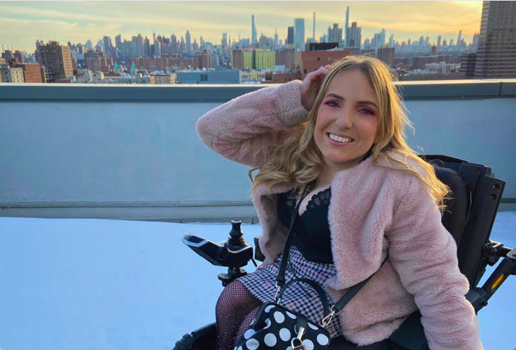 White woman with long blonde hair in a wheelchair smiling at the camera on a rooftop overlooking NYC
