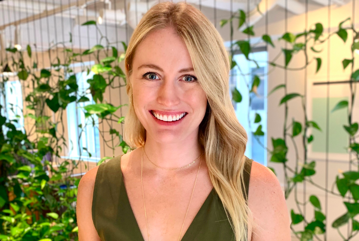White girl with blonde hair looking at the camera & smiling; She wear a sleeveless v-neck army green jumpsuit and stands in front of a background with leaves