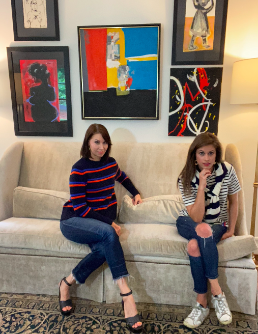 Two white women sitting on a couch with a gallery wall looking at the camera wearing striped tees