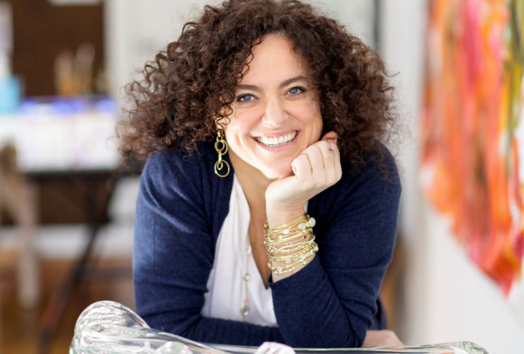 White woman with curly brown hair looking at the camera & smiling with her head leaning on her hand