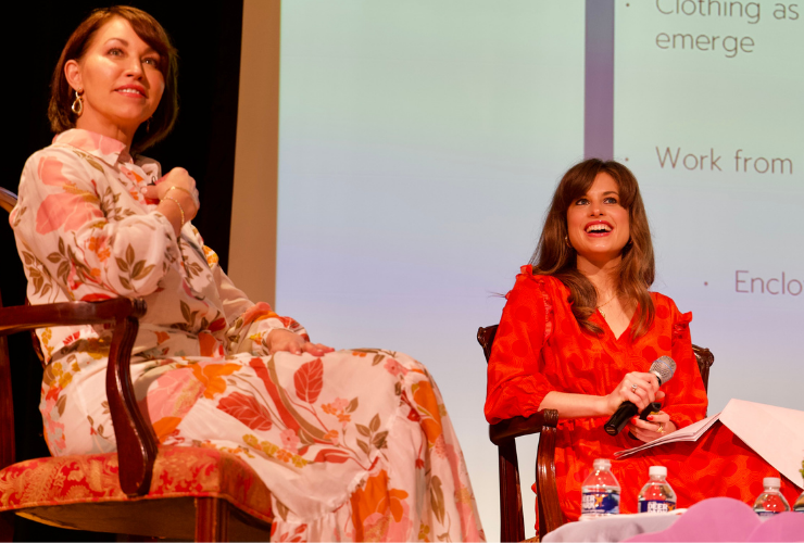 Two white women with brown hair wearing orange printed dresses sitting on stage holding microphones