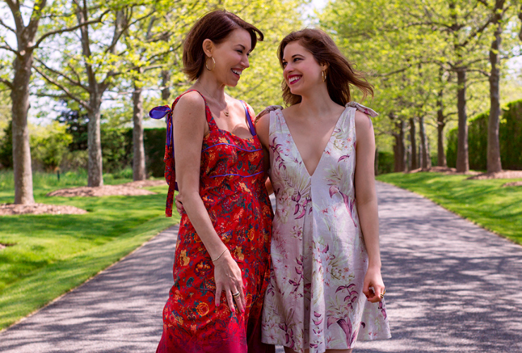 Two women standing on a road surrounded by trees with dappled sunlight; both have brown hair & wear printed dresses