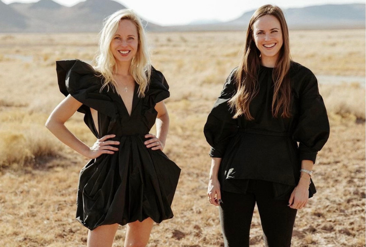 Two white women wearing black standing & smiling at the camera on a Texas ranch
