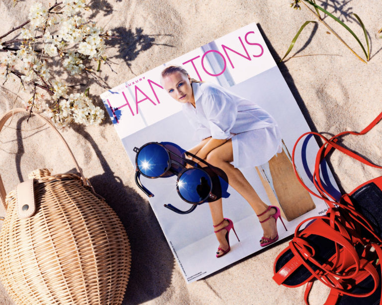 Flat lay of a straw bag, Hamptons Magazine, sunglasses & red sandals on sand